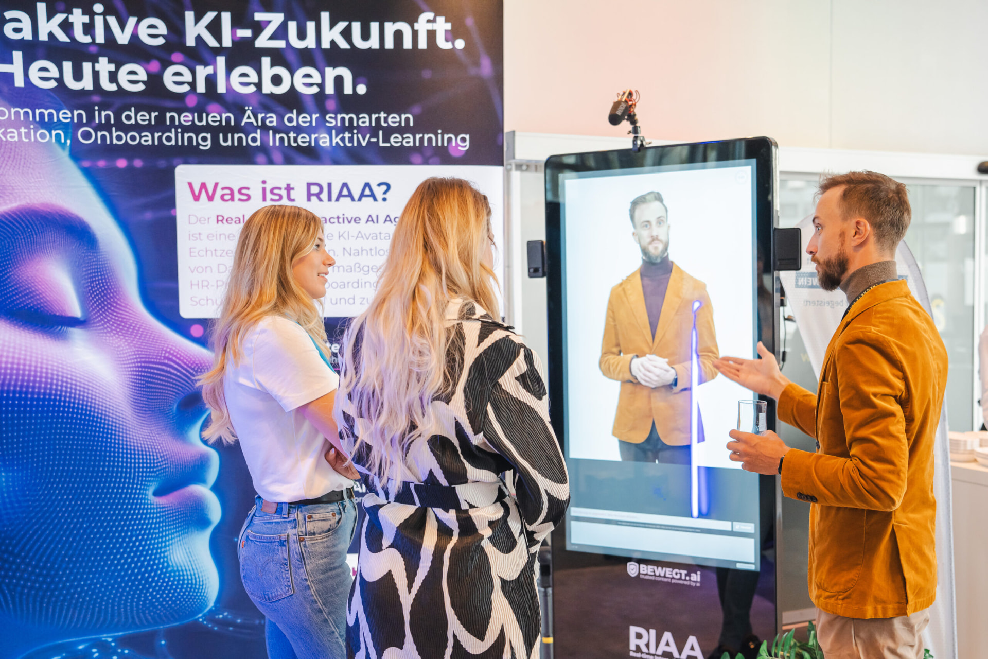 Besucherinnen im Gespräch an einem Bildschirm mit KI-Avatar auf einer Messe